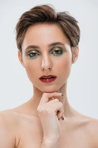 Портрет молодой женщины с ярким макияжем касающимся подбородка и смотрящей на камеру, изолированную на сером — стоковое фото