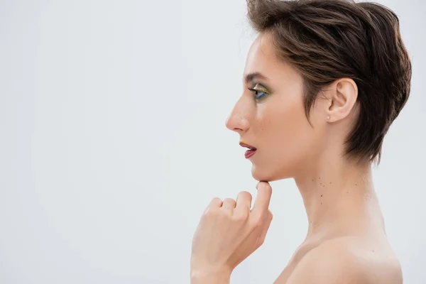 Seitenansicht einer jungen Frau mit hellem Make-up, die ihr Kinn isoliert auf grau berührt — Stockfoto