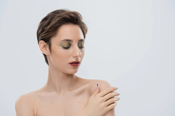 Ritratto di giovane donna con trucco luminoso e capelli corti che toccano la spalla nuda e guardano la fotocamera isolata sul grigio — Foto stock