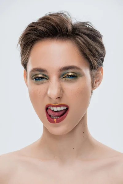 Portrait of emotional young woman with shiny makeup and short hair sticking out tongue isolated on grey — Stock Photo