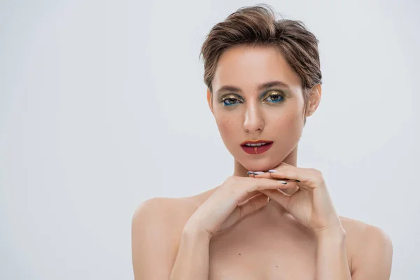 Junge Frau mit schimmerndem Augen-Make-up und kurzen Haaren blickt vereinzelt in die Kamera auf grau — Stockfoto