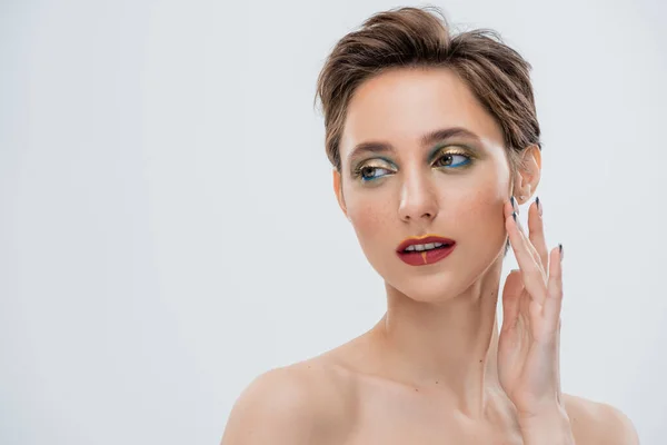 Junge Frau mit schimmerndem Augen-Make-up und kurzen Haaren, die die Wange berühren, isoliert auf grau — Stockfoto