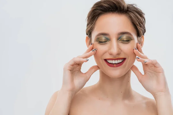 Jeune femme satisfaite avec un maquillage des yeux chatoyant touchant les joues isolées sur gris — Photo de stock