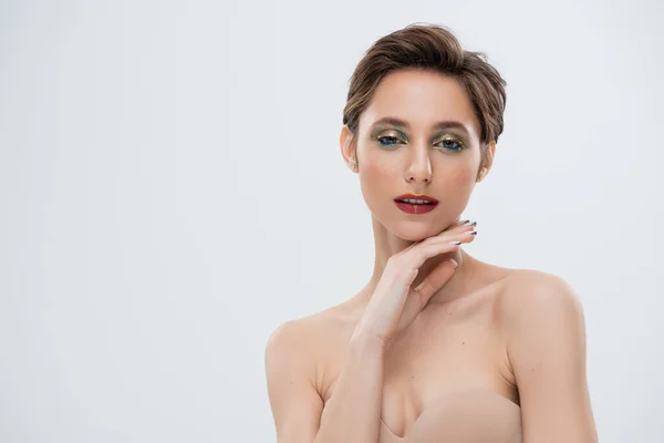 Hübsche junge Frau mit schimmerndem Augen-Make-up, das das Kinn isoliert auf grau berührt — Stockfoto