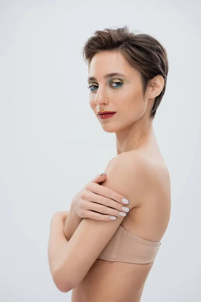 Hübsche junge Frau mit schimmerndem Augen-Make-up in trägerlosem BH isoliert auf grau — Stockfoto