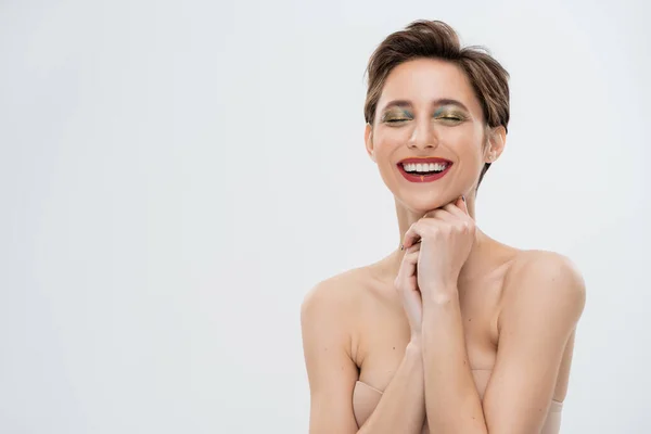 Freudige junge Frau mit schimmerndem Augen-Make-up im trägerlosen BH isoliert auf grau — Stockfoto