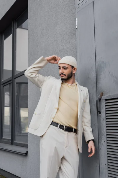 Hombre homosexual de moda en traje beige y sombrero mirando hacia otro lado cerca del edificio al aire libre - foto de stock