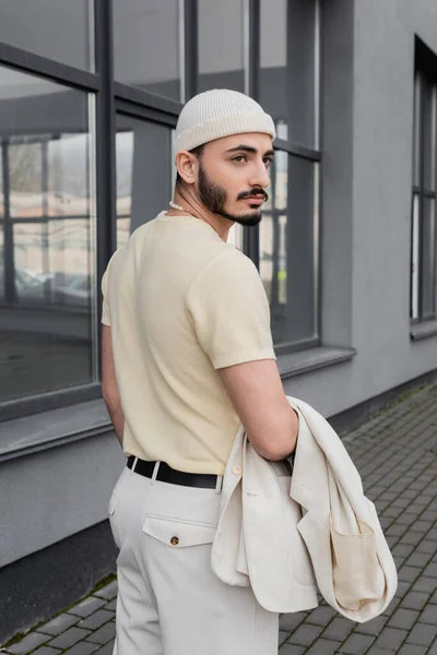 Модний гей-чоловік у капелюсі тримає піджак біля будівництва на відкритому повітрі — стокове фото
