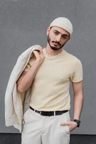 Retrato de hombre gay de moda en sombrero sosteniendo chaqueta y posando al aire libre - foto de stock