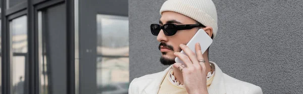 Trendiger schwuler Mann mit Sonnenbrille und Hut, der auf dem Handy in der Nähe von Gebäuden im Freien spricht, Banner — Stockfoto