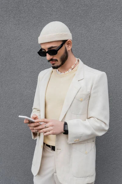 Модний гей-чоловік в сонцезахисних окулярах використовує смартфон біля стіни на відкритому повітрі — стокове фото