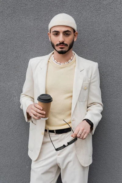 Elegante gay homem no terno segurando café para ir e óculos de sol perto de parede ao ar livre — Fotografia de Stock