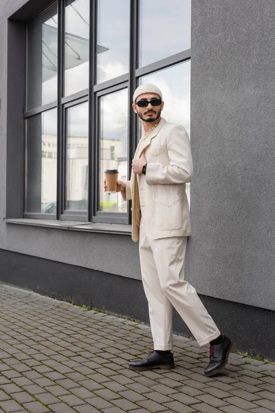 Гей в стильном костюме и солнечных очках держит кофе, чтобы выйти на городскую улицу — стоковое фото