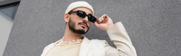 Vista basso angolo di uomo gay alla moda in cappello toccare occhiali da sole vicino edificio sulla strada urbana, banner — Foto stock