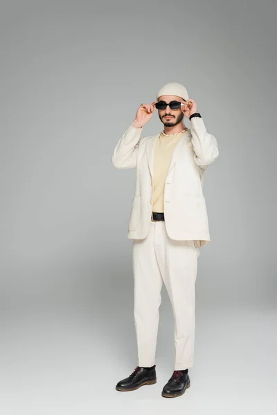 Piena lunghezza di alla moda gay uomo in beige vestito e cappello indossare occhiali da sole su sfondo grigio — Foto stock
