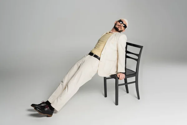 Добре одягнений гей чоловік в сонцезахисних окулярах позує зі стільцем на сірому фоні — стокове фото