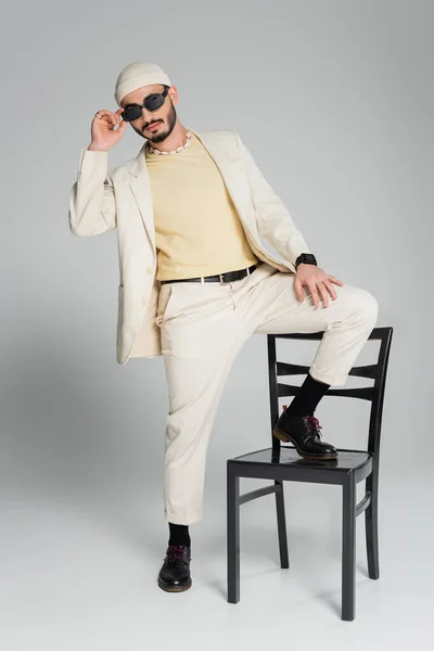 Hombre gay de moda en traje y gafas de sol posando con silla sobre fondo gris - foto de stock