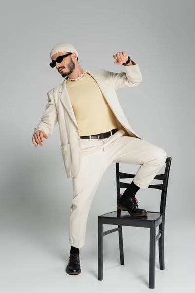 Trendy gay man in suit and sunglasses posing near chair on grey background — Stock Photo