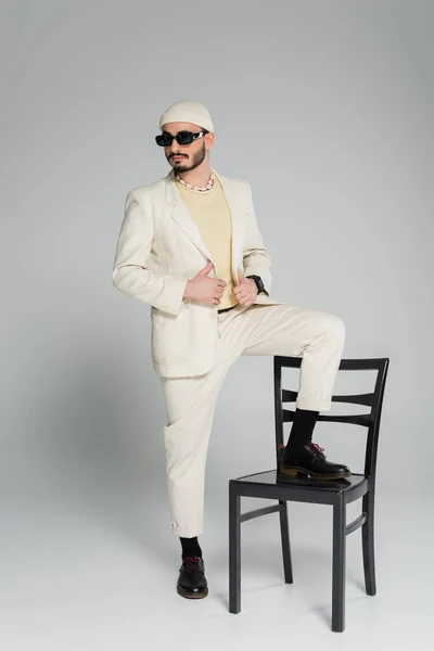 Hombre homosexual de moda en gafas de sol posando cerca de la silla sobre fondo gris - foto de stock