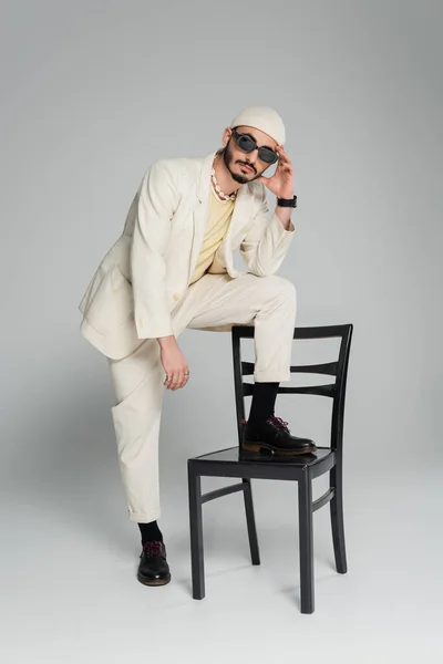 Модный гомосексуалист в костюме и шляпе позирует возле стула на сером фоне — стоковое фото