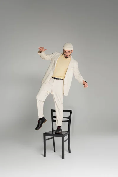Stylish gay man in beige suit and hat standing on chair on grey background — Stock Photo