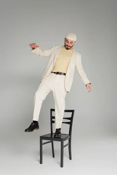 Модный гомосексуалист в костюме и бежевая шляпа, стоящая на стуле на сером фоне — стоковое фото
