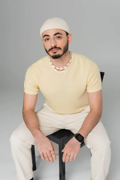 Модний гей чоловік в капелюсі дивиться на камеру, сидячи на стільці на сірому фоні — стокове фото