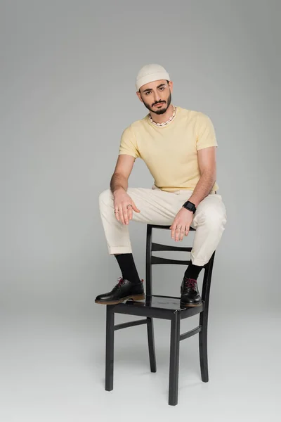 Bärtiger schwuler Mann mit Hut sitzt auf Stuhl auf grauem Hintergrund — Stockfoto