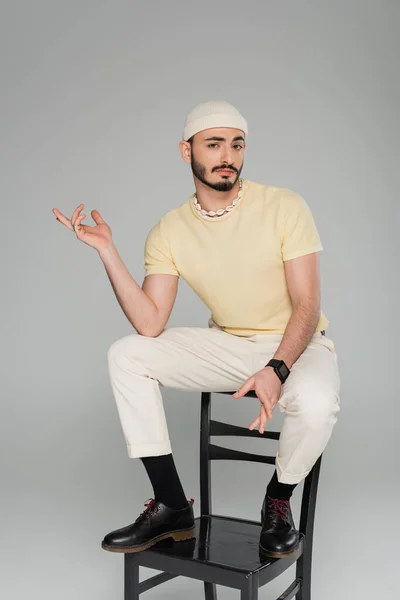 Confuso gay homem no chapéu sentado no cadeira isolado no cinza — Fotografia de Stock