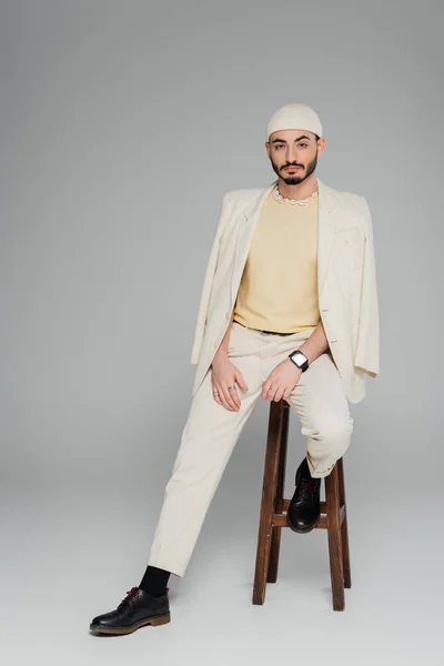 Homme gay à la mode en costume beige et chapeau assis sur la chaise sur fond gris — Photo de stock