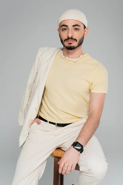 Ritratto di uomo gay alla moda in giacca e cappello in posa sulla sedia isolata su grigio — Foto stock
