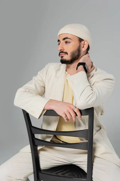 Barbuto gay uomo in beige cappello e vestito guardando lontano mentre seduto su sedia isolato su grigio — Foto stock