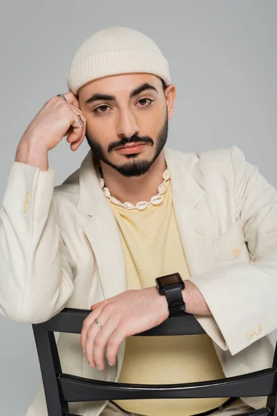 Ritratto di uomo omosessuale alla moda in cappello e giacca seduto sulla sedia isolato su grigio — Foto stock