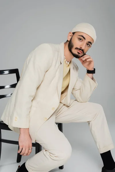 Модный гомосексуалист в шляпе и бежевом костюме позирует на стуле на сером фоне — стоковое фото