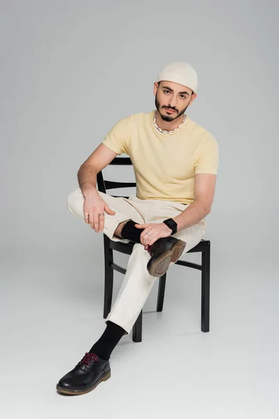 Na moda gay homem no chapéu e concha colar sentado no cadeira no cinza fundo — Fotografia de Stock
