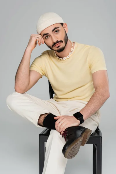 Портрет стильного молодого гея в шляпе, сидящего на стуле изолированного на сером — стоковое фото