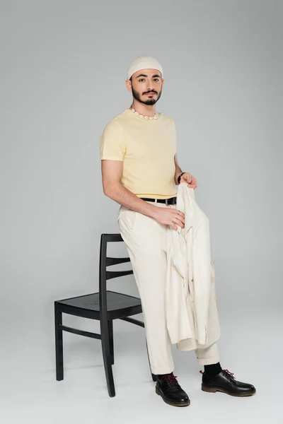 Повна довжина модного гомосексуального чоловіка в капелюсі тримає піджак біля стільця на сірому фоні — стокове фото