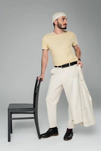 Full length of well dressed homosexual man holding jacket near chair on grey background — Stock Photo