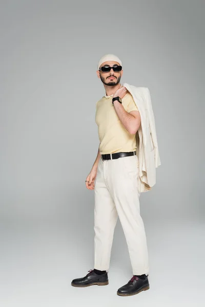 Elegante gay hombre en gafas de sol celebración chaqueta mientras de pie en gris fondo - foto de stock