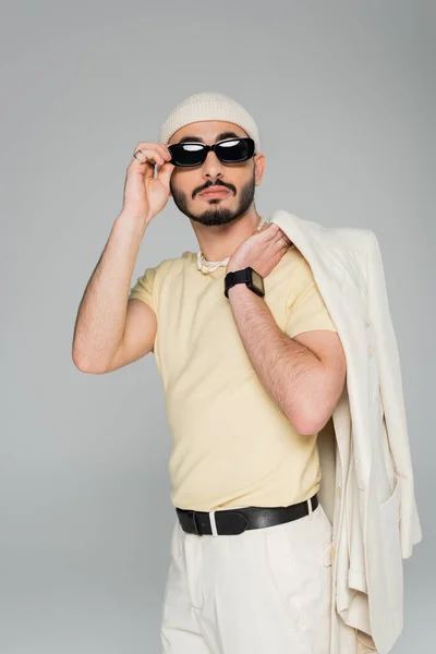Na moda gay homem no chapéu segurando jaqueta e tocando sol óculos isolado no cinza — Fotografia de Stock