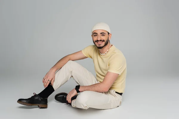 Cheerful gay man in hat looking at camera while sitting on grey background — Stock Photo