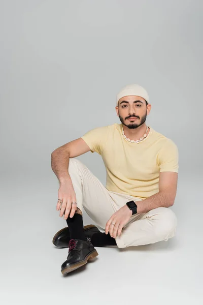 Бородатый гомосексуалист в шляпе смотрит в камеру, сидя на сером фоне — стоковое фото