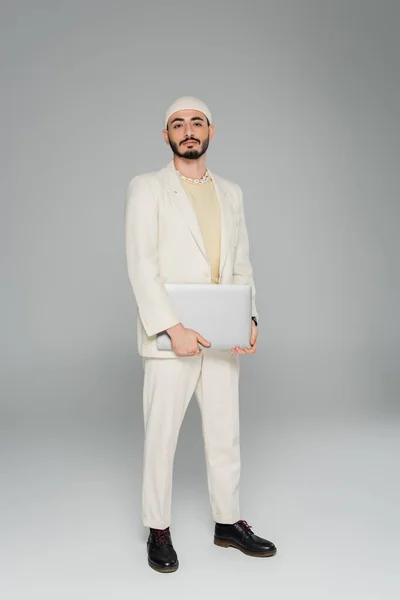 Stylish gay man in beige suit holding laptop on grey background — Stock Photo