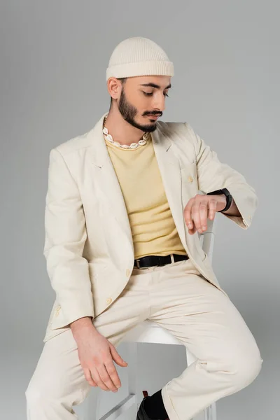 Стильний гей чоловік у костюмі дивиться на розумний годинник, сидячи на стільці ізольовано на сірому — стокове фото