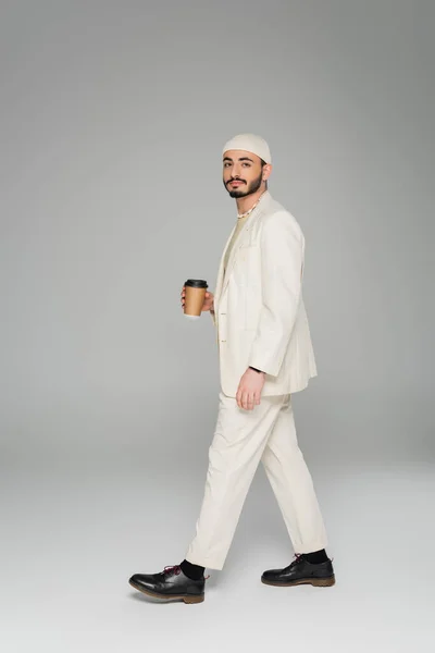 Stylish gay man in suit holding coffee to go and walking on grey background — Stock Photo