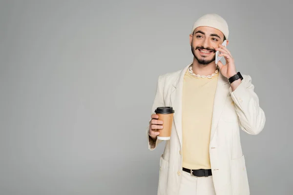 Улыбающийся гомосексуалист в костюме разговаривает по смартфону и держит кофе, чтобы быть изолированным от серого — стоковое фото