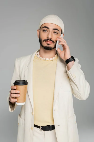 Хорошо одетый гей в костюме держит бумажную чашку и разговаривает на смартфоне изолирован на серый — стоковое фото