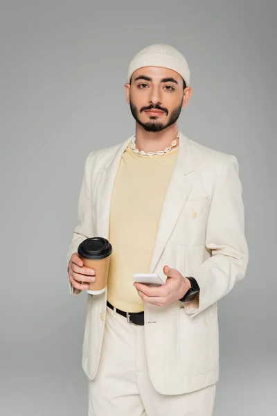 Стильний гомосексуальний чоловік у костюмі тримає смартфон і каву, щоб ізолюватись на сірому — стокове фото