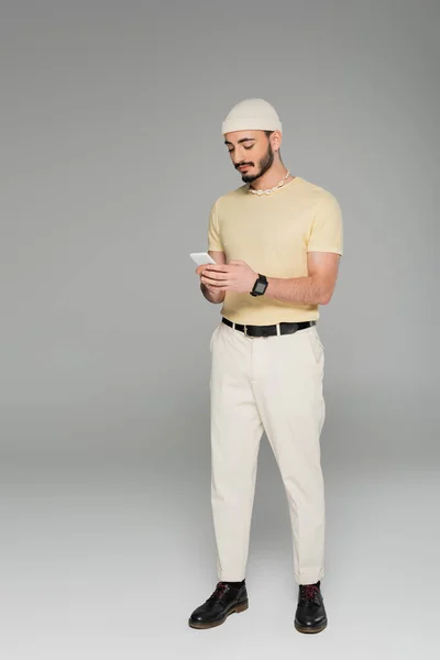 Trendy gay man in hat using cellphone while standing on grey background — Stock Photo