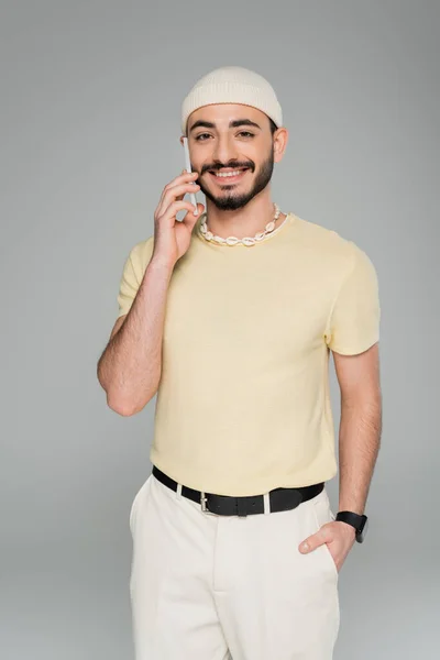 Усміхнений гей чоловік в капелюсі говорить на смартфоні і позує ізольовано на сірому — стокове фото